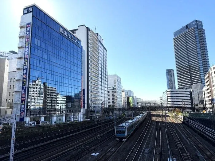 青いガラス張りのビルや高層ビルが立ち並ぶ都市の風景。手前には複数の鉄道の線路が広がり、一編成の電車が走っている。