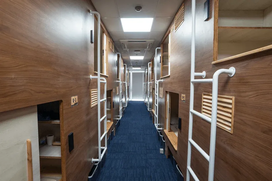 木目の壁と青いカーペットが特徴的な、カプセルホテルの廊下の様子。両側に複数のカプセルユニットが並び、上段には梯子でアクセスする。コンパクトながら機能的な宿泊空間が広がっています。