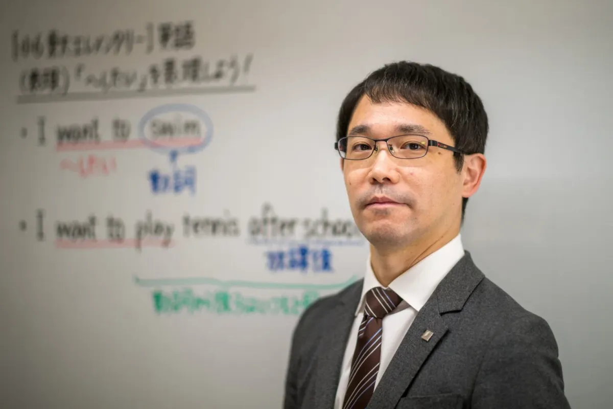 スーツを着た男性が、英語の「I want to」の表現について書かれたホワイトボードの前に立っています。