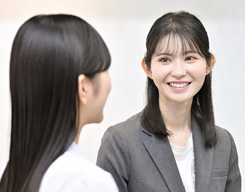 グレーのジャケットを着た女性が、もう一人の女性と向き合い、明るい笑顔を見せています。和やかな雰囲気の会話の様子です。