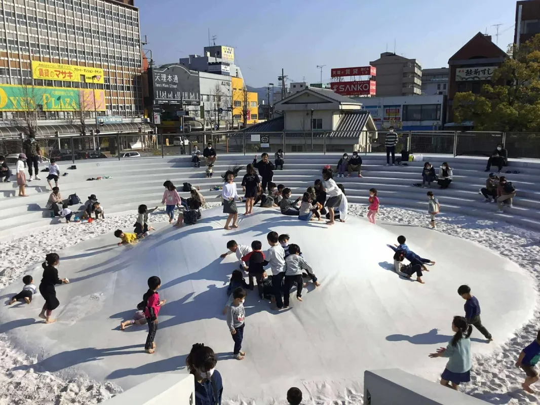 多くの子供たちが白いドーム型の遊具と砂場で遊んでいる屋外の広場の様子