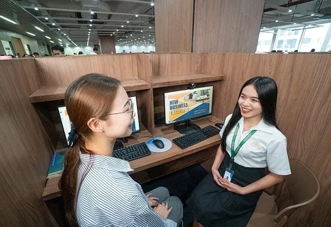 モダンな学習スペースで、2人の女性がパソコンを囲んで会話しています。片方の女性は笑顔で、画面には「NEW BUSINESS ENGLISH」と表示されています。