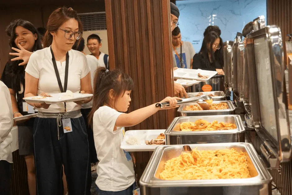 多くの人々がビュッフェラインに並び、温かい料理を各自の皿に盛り付けている様子。子供も大人も楽しそうに食事を選んでいます。