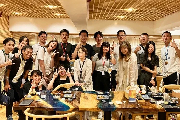 多様な人々がレストランで食事会を楽しんでいる集合写真です。参加者たちは皆笑顔で、ピースサインやサムズアップをしており、和やかな雰囲気が伝わってきます。