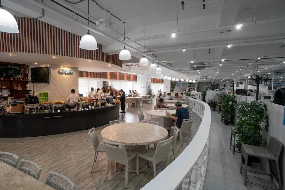 広々としたモダンなカフェテリアで、人々が食事を楽しんでいます。ビュッフェカウンターには料理が並び、スタッフがサービスを提供しており、明るく活気のあるダイニング空間が広がっています。