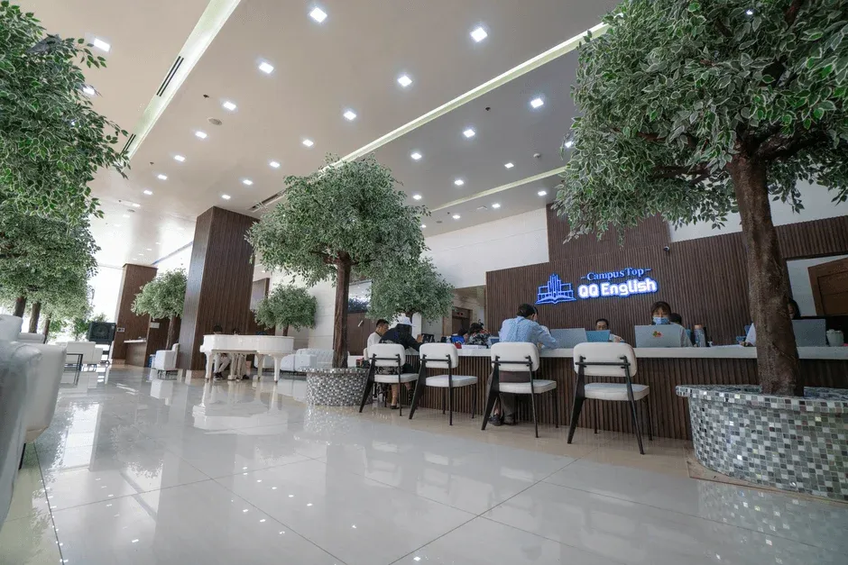 英語学校の広々としたモダンな受付または学習スペース。天井が高く、人工樹木が配置されており、明るく清潔感のある雰囲気です。数人がラップトップで作業しており、奥には白いグランドピアノも見えます。