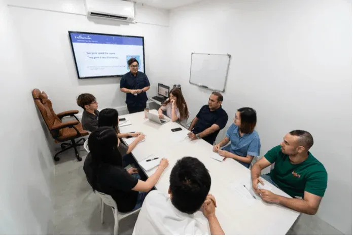 会議室で男女数人がテーブルを囲み、一人がスクリーンを使ってプレゼンテーションをしています。参加者たちは熱心に耳を傾け、活発な議論が行われているビジネスミーティングの様子です。