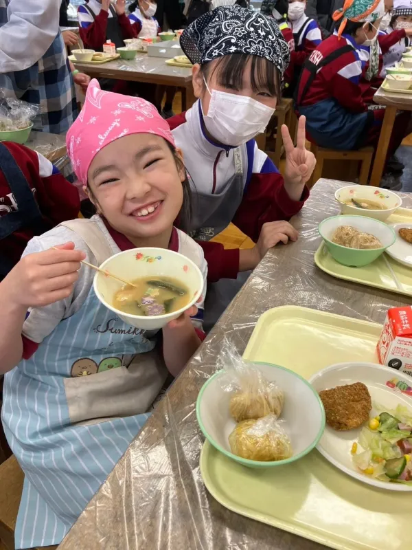バンダナとエプロンをつけた小学生の女の子二人が、学校の給食を笑顔で楽しんでいる