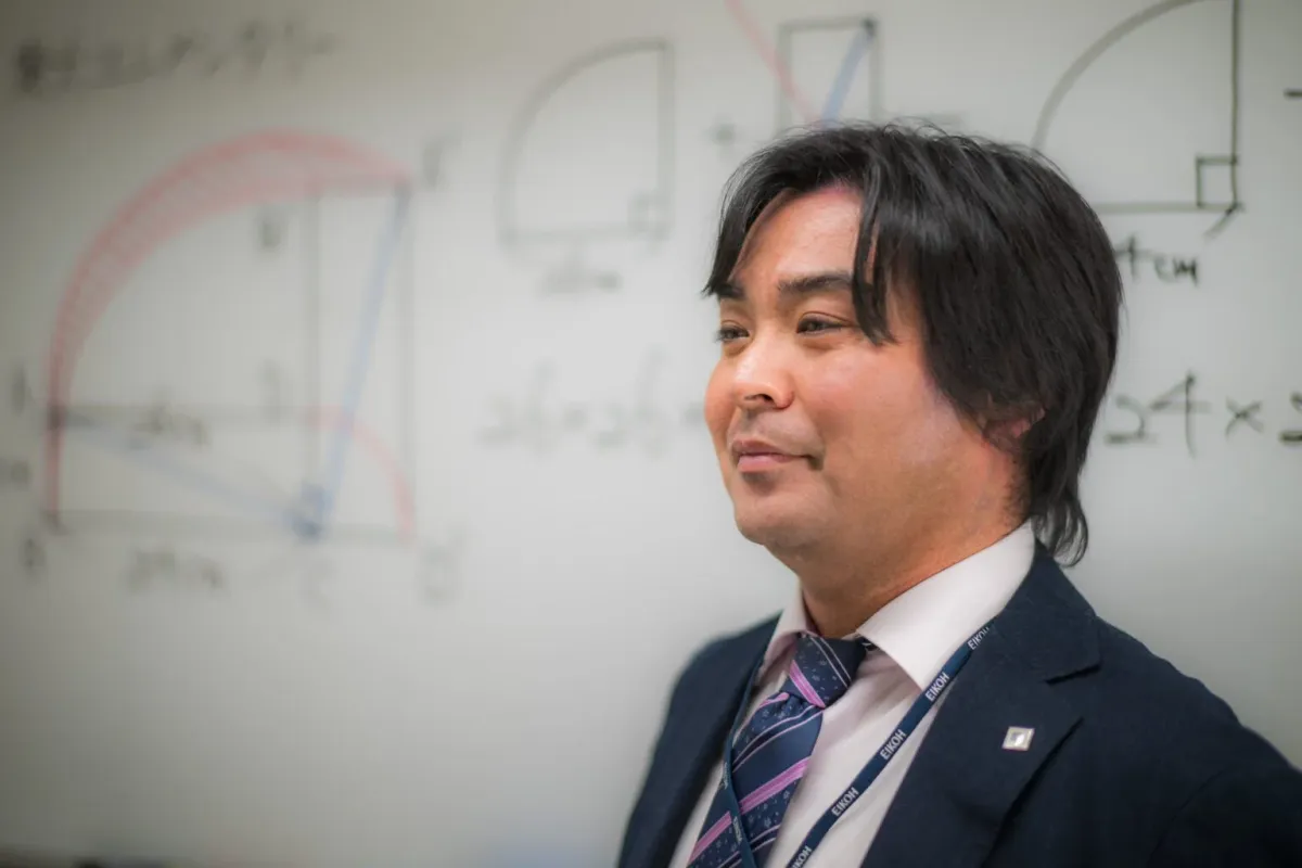 EIKOHのストラップをつけた男性が、数学の図形や数式が書かれたホワイトボードの前で横を向いています。