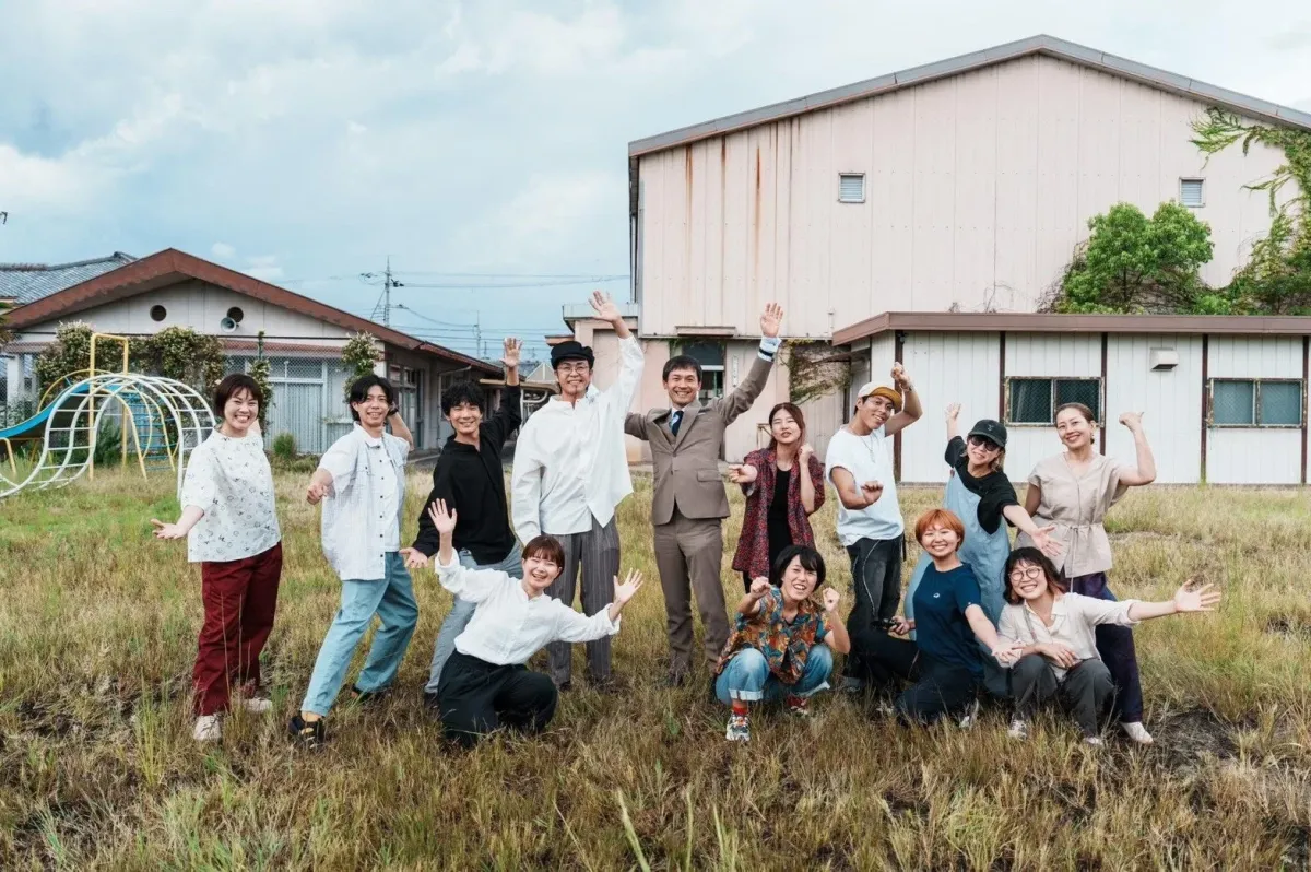 広がる草原で約15人の男女が笑顔で手を振ったりポーズを取ったりしている集合写真