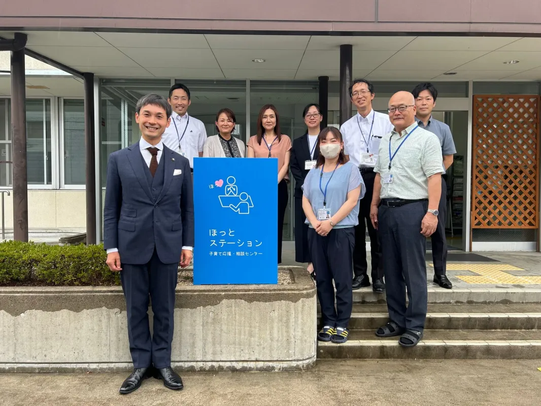 施設の前で、笑顔のスタッフ9人が並んでポーズをとっています