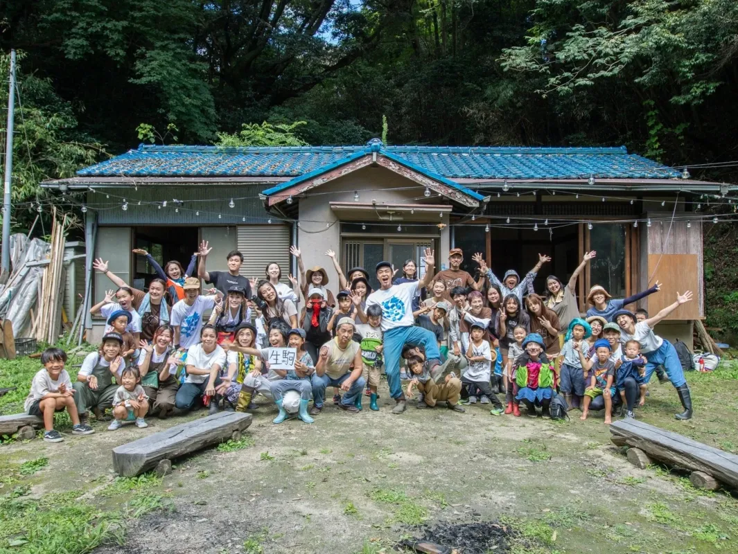青い瓦屋根の古民家の前で、老若男女が大勢で笑顔で手を振っている集合写真