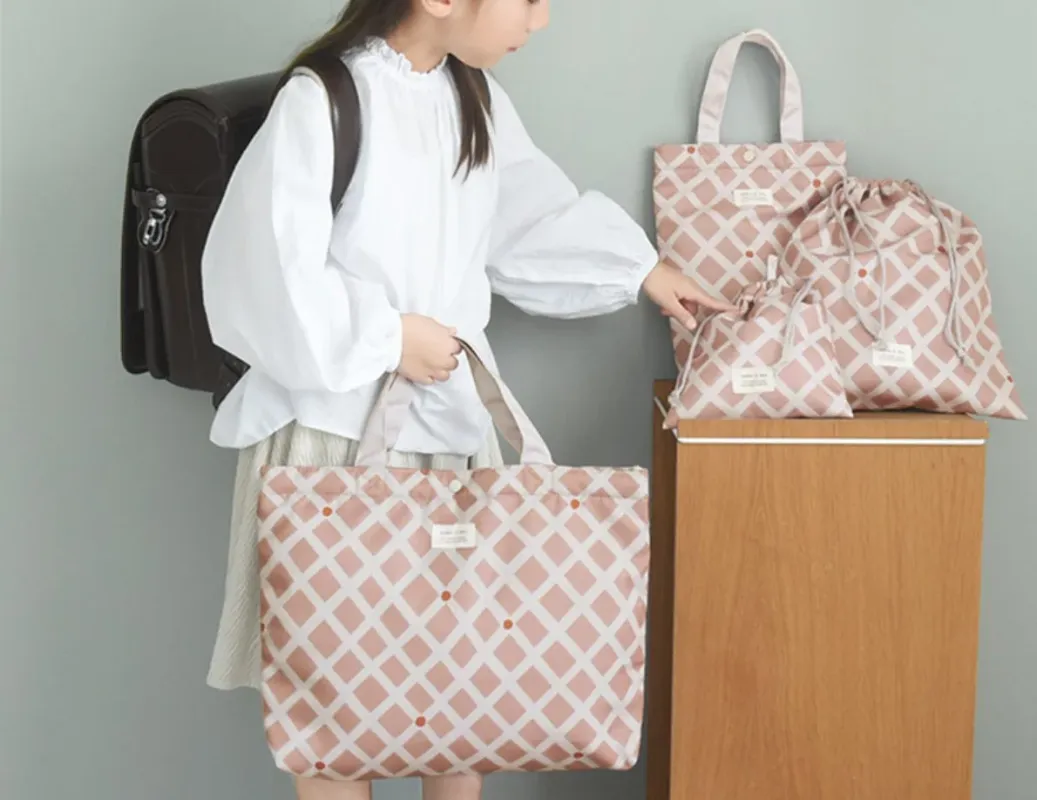 ランドセルを背負った少女が、ピンクのダイヤ柄のトートバッグや巾着袋などの通学グッズに囲まれている様子。新学期の準備を思わせる可愛らしい一枚です。