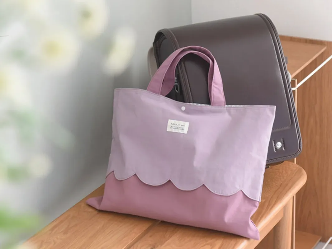 木製のベンチに置かれた、波型のデザインが特徴的なラベンダーとピンクのツートンカラーのトートバッグです。その奥には茶色のランドセルが見えます。新学期や通学準備を思わせる、可愛らしい雰囲気の写真です。