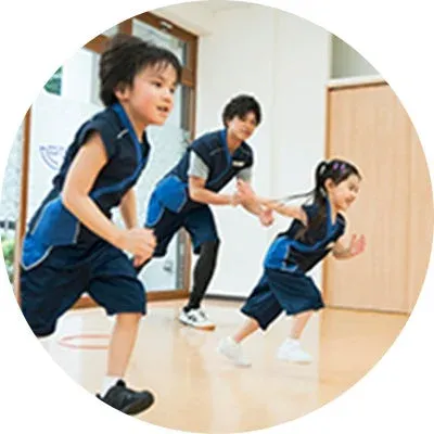 子供たちがトレーナーの指導のもと、室内で楽しそうに運動している様子です。青と黒のトレーニングウェアを着用し、活発に体を動かしています。