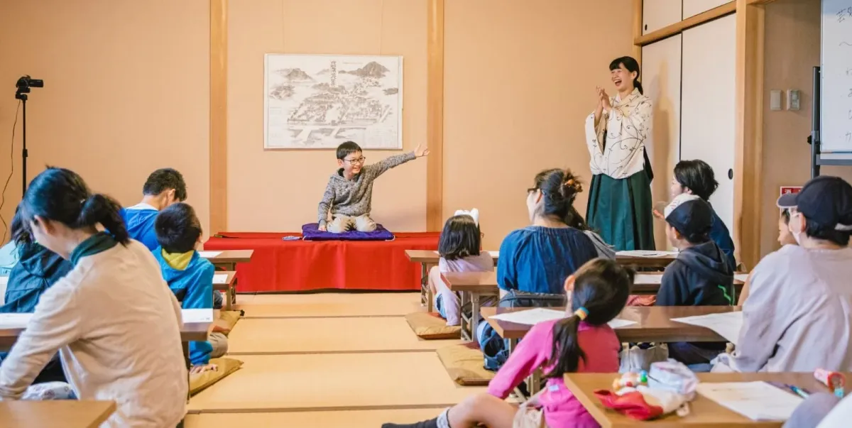 和室で子供たちが集まり、一人の少年が発表している様子。和服を着た女性が拍手で応援し、他の参加者も熱心に見守っている