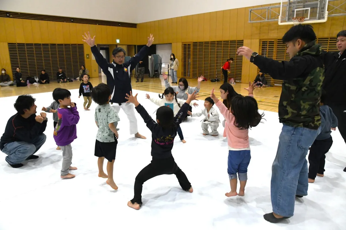 体育館の白いマットの上で、子供たちが大人と一緒に運動している