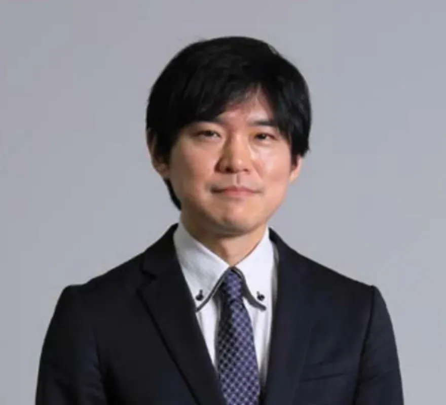 黒髪のアジア人男性が、ダークスーツに白いシャツ、柄物のネクタイを着用し、カメラをまっすぐ見つめている上半身のポートレート写真です