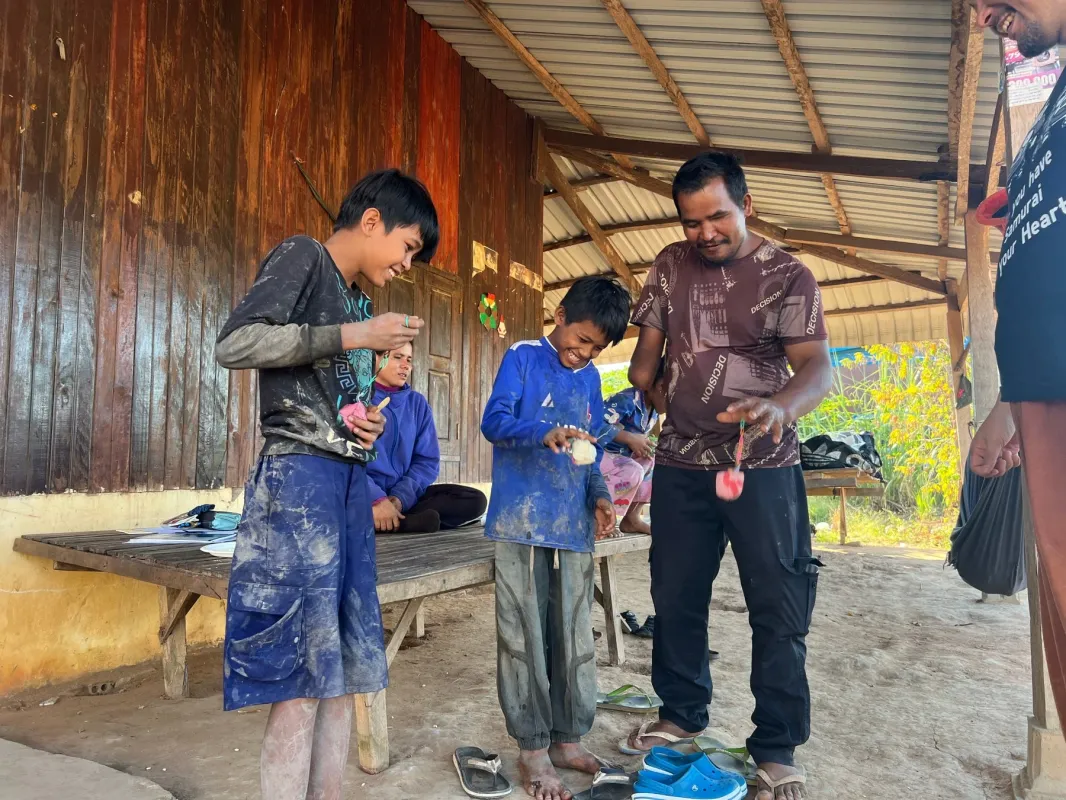 屋外の質素な家屋の前で、大人と子供たちが集まり、笑顔で交流している様子が捉えられています。男性が子供たちに何かを教えているようで、皆楽しげに手元の作業に集中しています。