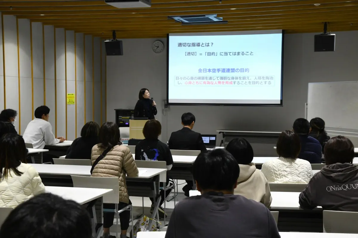 教室で、一人の女性がマイクを持ってプロジェクタースクリーンに映し出された資料を使い、大勢の聴衆にプレゼンテーションを行っている