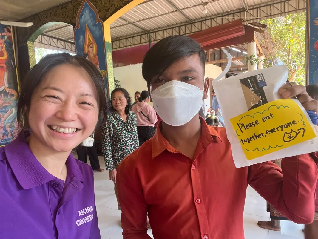 笑顔の女性とマスクの男性が、お菓子「はんなりチップス」と「皆で一緒に食べましょう」というメッセージの紙を持つ写真