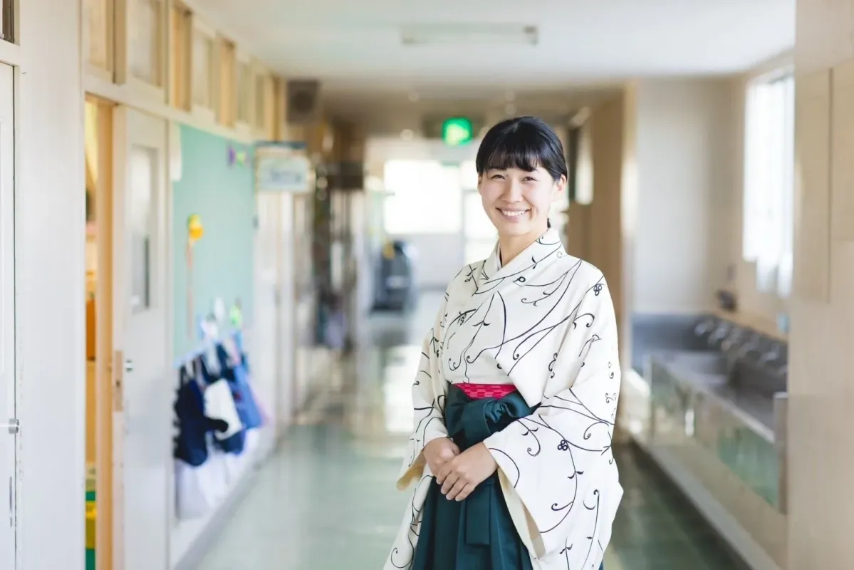 学校の廊下で、和服と袴を着用した笑顔の日本人女性が立っています