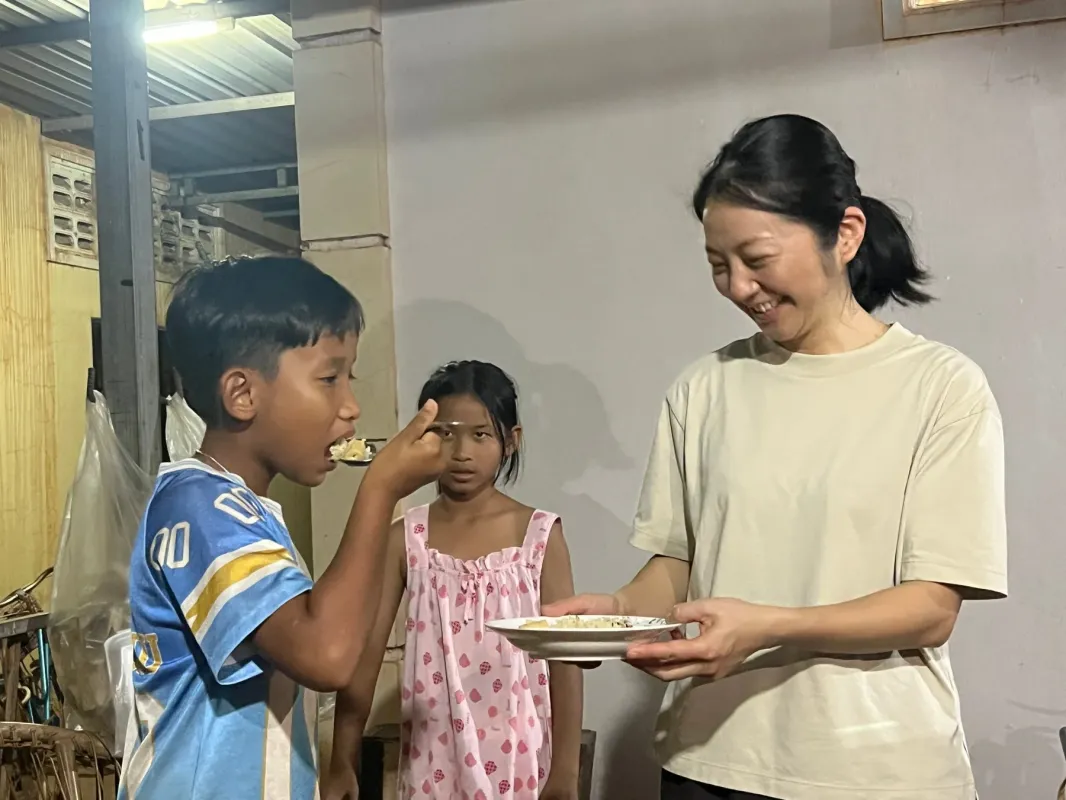 笑顔の女性が男の子にスプーンで食事を与えています。後ろにはもう一人の女の子が立っており、温かい雰囲気の中で食事が分かち合われている様子が伝わります。支援や交流の瞬間を捉えた心温まる一枚です。