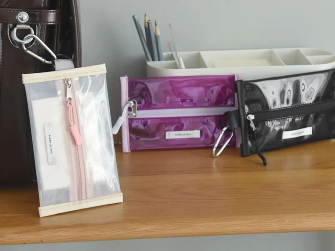 木製のテーブルに、クリア、紫、黒の3色の透明ポーチが並べられています。それぞれカラビナ付きで、小物や文房具の整理に便利なデザインです。背景には文房具やバッグが見えます。