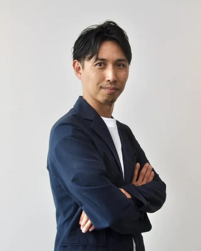 白いTシャツの上に紺色のブレザーを羽織った男性が、腕を組んでカメラを見つめているポートレート写真です。穏やかな表情で、ビジネスシーンやプロフィール写真に適しています。