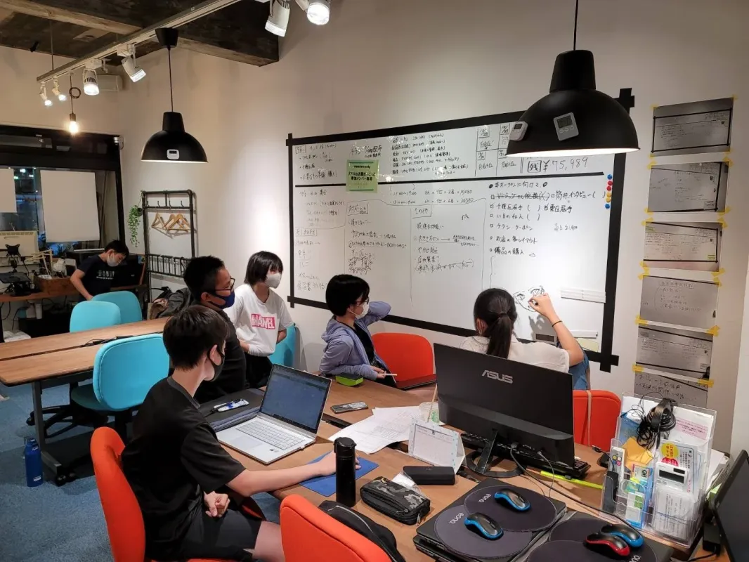 オフィス空間で数人がホワイトボードを囲んで打ち合わせをしている様子。一人がホワイトボードに書き込み、他のメンバーはそれを見たり、自身のPCで作業したりしている。活気ある共同作業の場面。