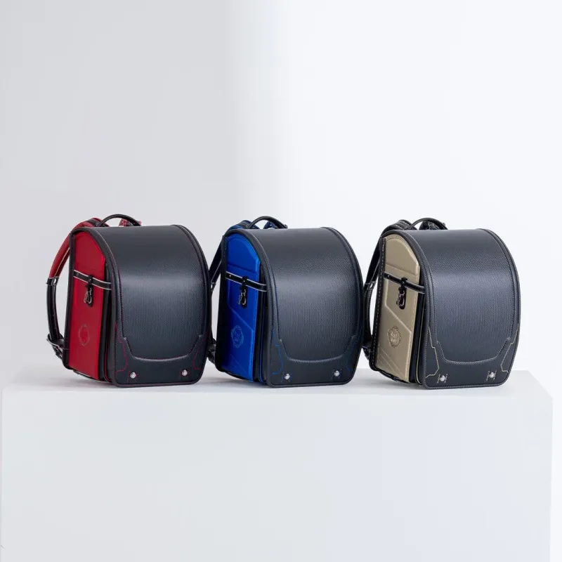 黒を基調とした赤、青、ベージュの3色のランドセルが白い台の上に並べられています。