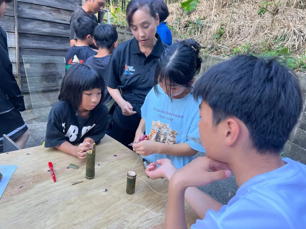 屋外の木製テーブルで、子供たちが大人の指導のもと竹細工のようなクラフト活動に集中している様子。教育的なワークショップの一場面です。