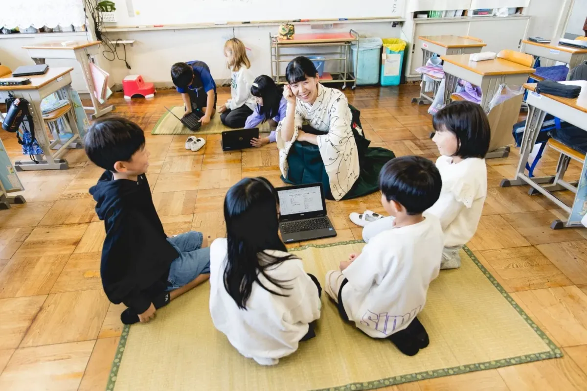 畳敷きの教室で、着物姿の女性教師が子供たちとノートパソコンを使ったグループ学習をしている様子