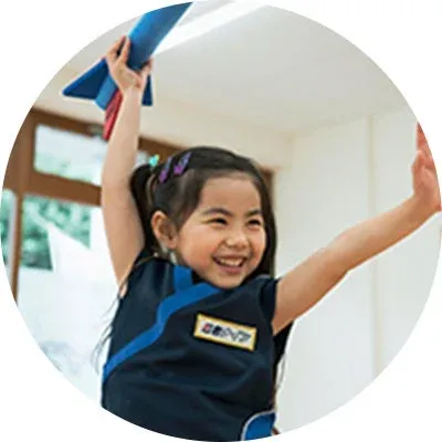 青い服を着た小さな女の子が、青いロケットのおもちゃを掲げて満面の笑みを浮かべている様子。楽しそうな雰囲気の屋内での一コマです。