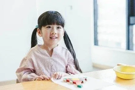 明るい室内で、ピンクの服を着たポニーテールの女の子が、カラフルなブロックで遊んでいます。