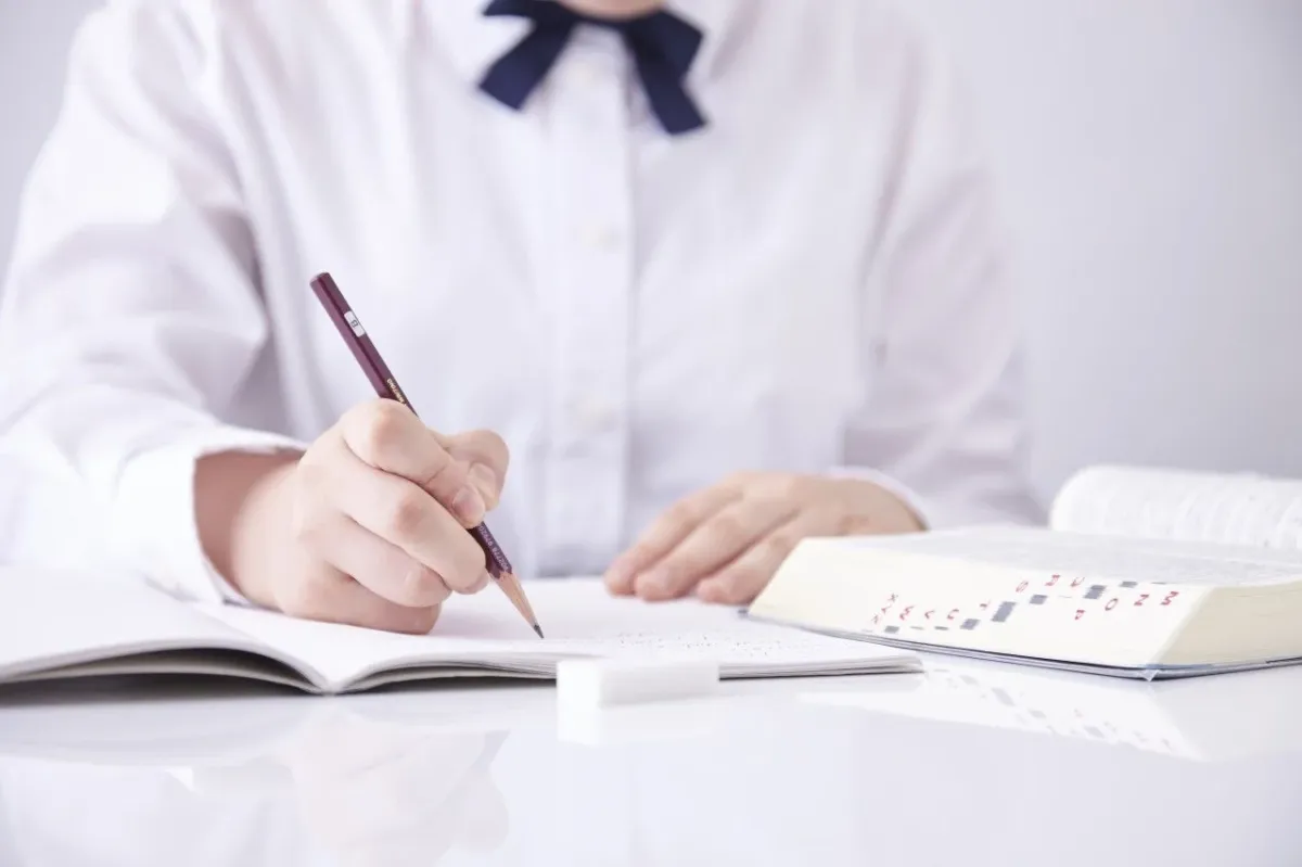 白いシャツにネイビーのリボンをつけた人物が、白い机の上で鉛筆を使いノートに書き物をしている様子です。隣には開かれた教科書と消しゴムがあり、勉強や学習に集中している場面を捉えています。