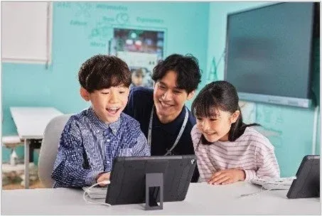 教室で先生と2人の子供たちがタブレット端末を囲んで楽しそうに学習している様子