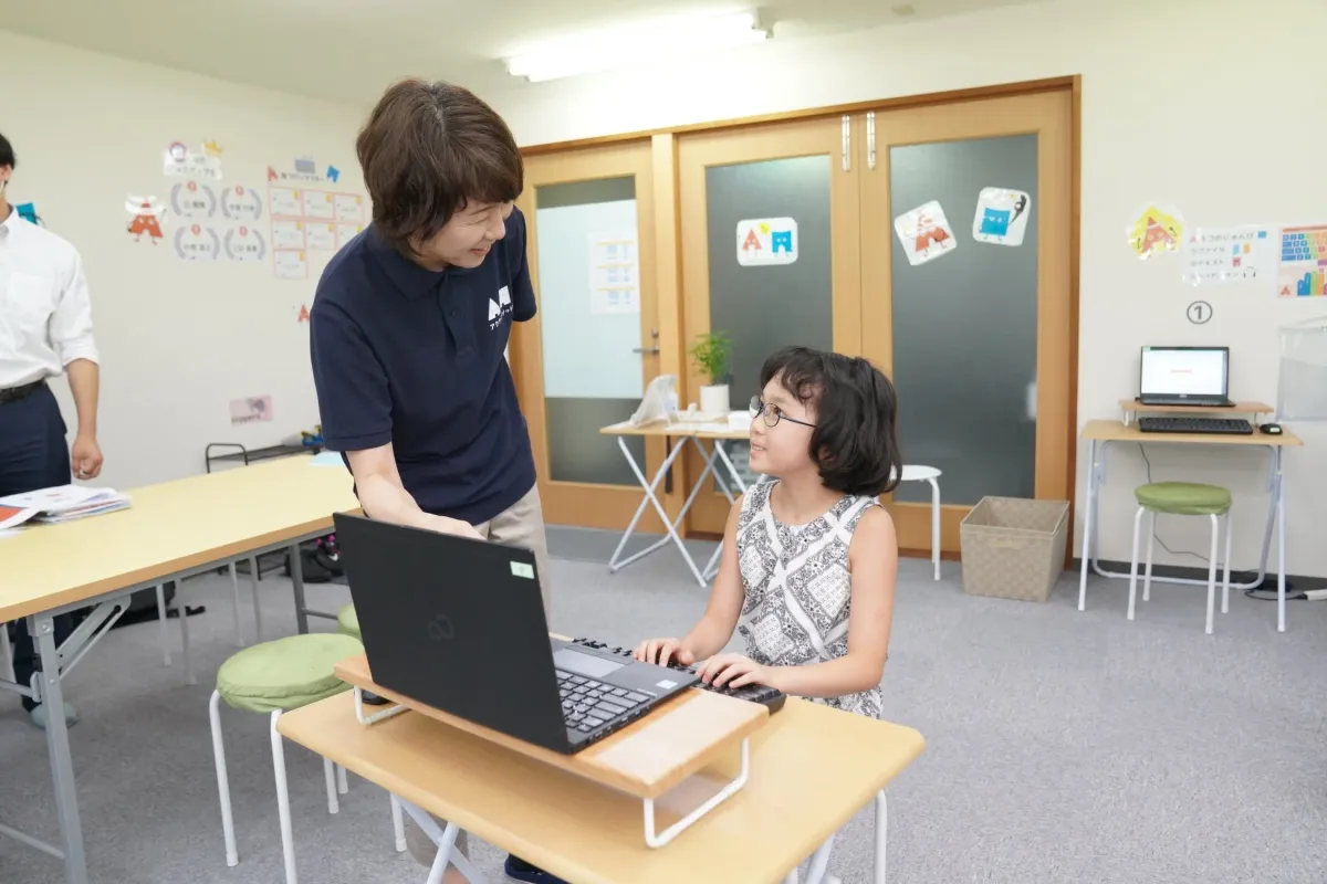 教室で女性の指導者がノートパソコンを使う少女に教えている様子
