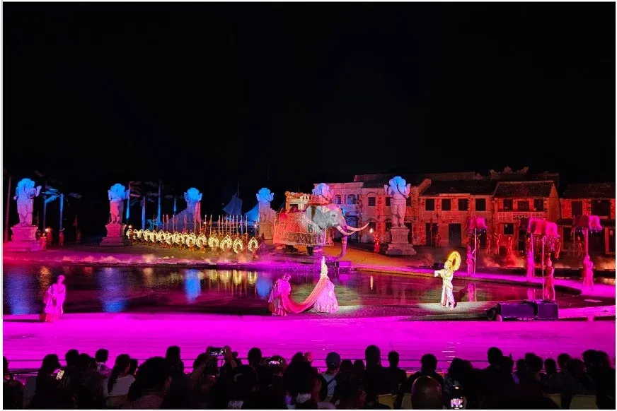 夜間に開催される大規模な屋外ショーの様子。伝統衣装のパフォーマー、装飾された象、歴史的な建物や像が鮮やかな光で照らされ、水面に反射する光が幻想的な雰囲気を醸し出している。観客がその壮大なスペクタクルを楽しんでいる。