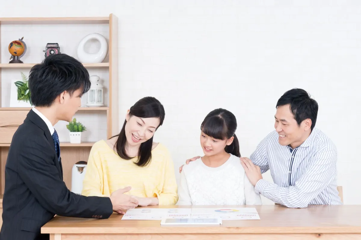 スーツ姿の男性が、家族(夫婦と娘)に対して資料やタブレットを使って何かを説明している場面です。家族は笑顔で話を聞いており、和やかな雰囲気で相談が進められている様子が伺えます。