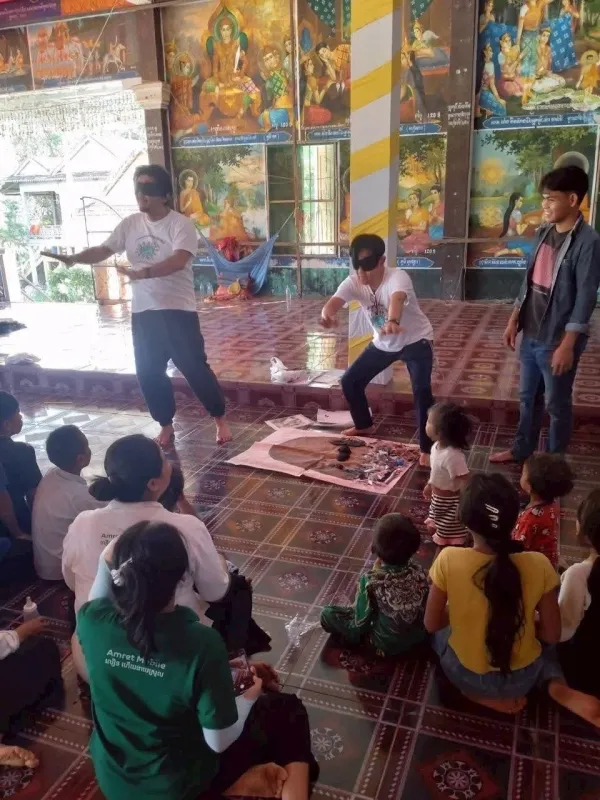 寺院のような屋内で目隠しゲームをする大人と見守る子どもたち