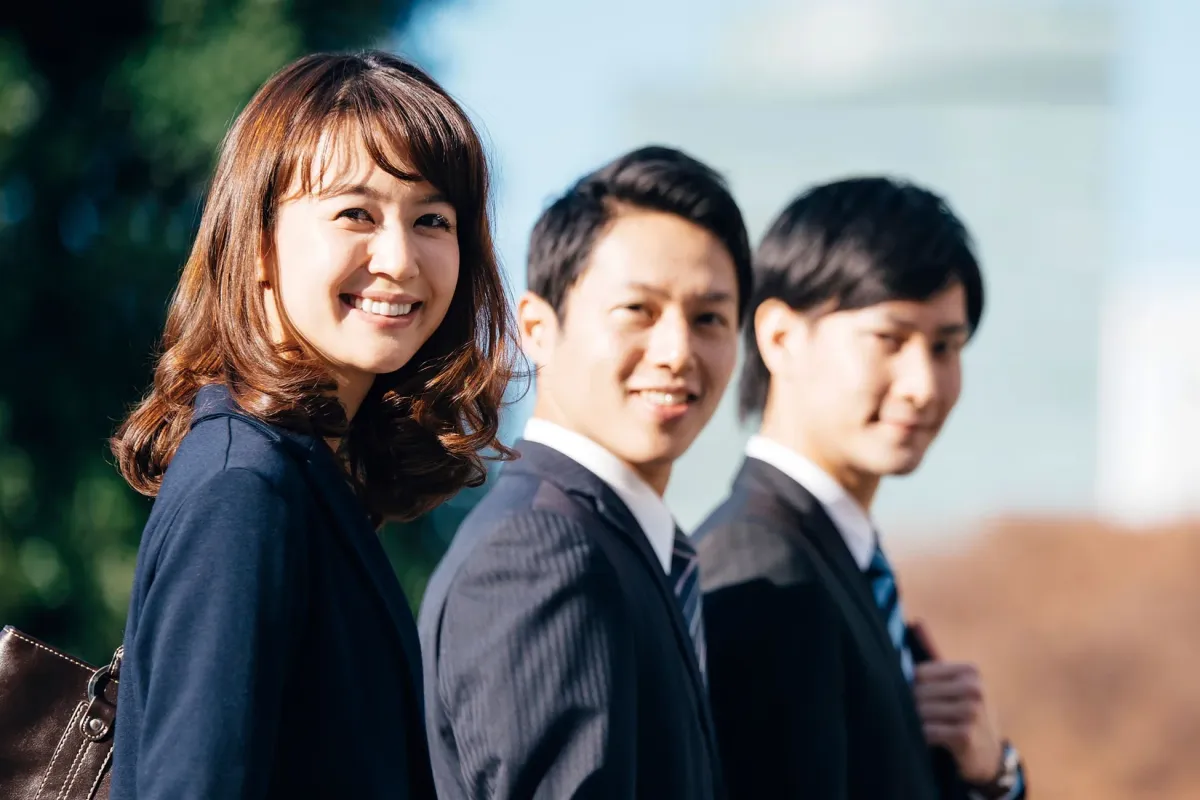 屋外で笑顔を見せる3人の若手ビジネスパーソン(女性1人、男性2人)の集合写真です。全員がカメラに向かって明るく微笑んでおり、フレッシュで親しみやすい印象を与えます。ビジネスシーンやチームワークを想起させる一枚です。