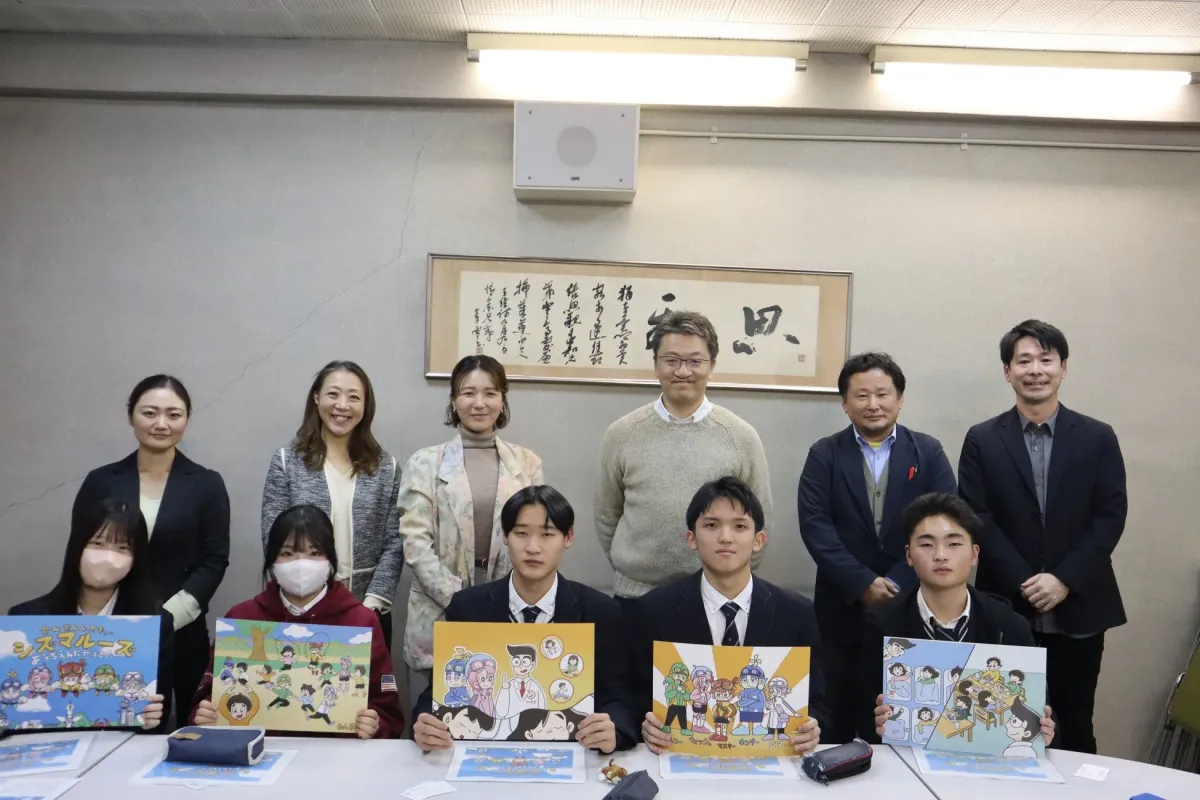 学生と大人が集まった集合写真。手前の学生たちは、がんや肝臓病に関するイラスト入りの教育資料を提示しており、健康教育プロジェクトの発表会の様子と見られる。