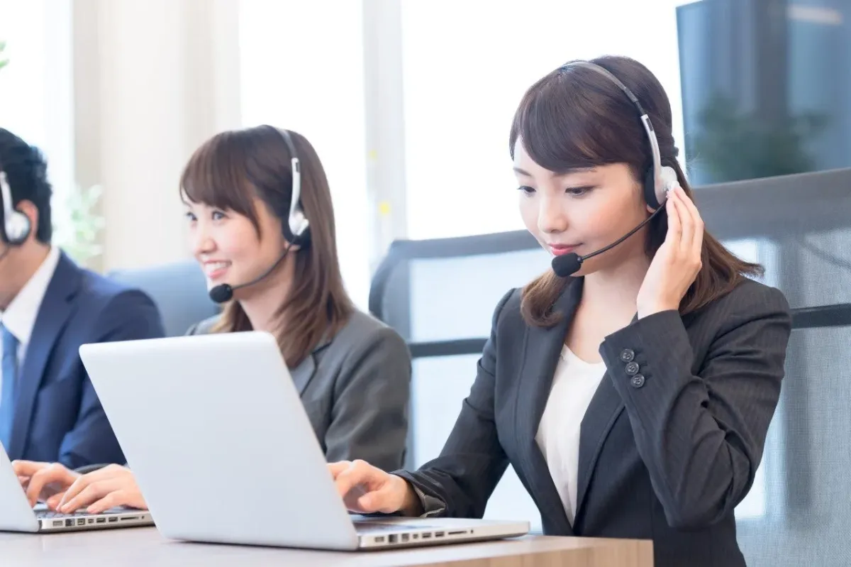 オフィスでヘッドセットを装着し、ノートパソコンを使用している3人のオペレーターが業務にあたっている様子です。手前の女性は真剣な表情で、奥の女性は笑顔で対応しています。