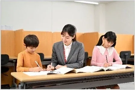 塾や学習塾のような場所で、女性の先生が小学生の男の子と女の子の学習をサポートしている様子