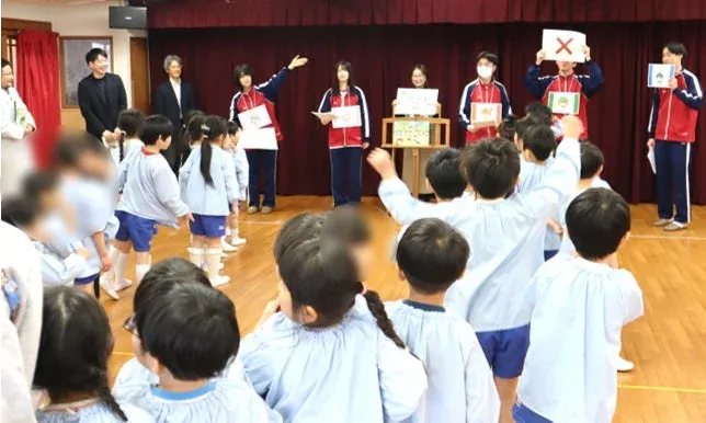 幼稚園か保育園のホールで、園児たちが大人たちによる教育的なイベントに参加している場面です。大人たちは絵や「X」マークのカードを提示し、園児たちは楽しそうに手を挙げています。