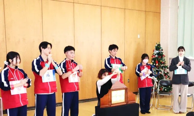 体育館のような場所で、複数の学生が何らかのイベントや発表を行っている様子です。赤いジャージを着た学生たちが中心におり、クリスマスツリーも飾られています。