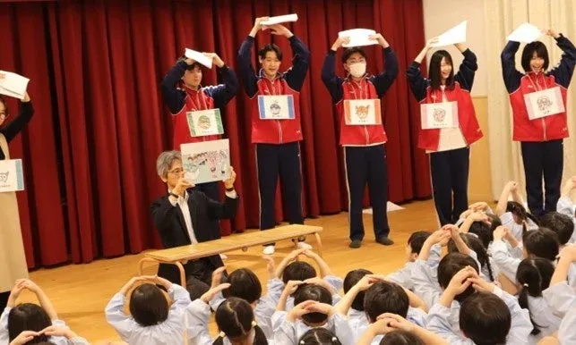 舞台上で学生たちが動物の名前が書かれたカードや絵本を使ってパフォーマンスを行っており、多くの園児たちが床に座ってそれを見ている様子。教育的なイベントや劇が開催されているようだ。