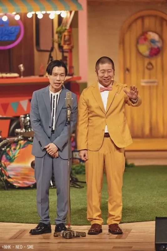 カラフルなステージで、スーツを着た男性2人が笑顔で立っています