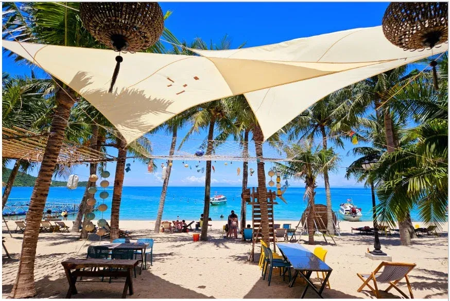 熱帯のビーチにあるリゾート施設で、白い砂浜にはヤシの木が立ち並び、日よけのタープの下にはテーブルと椅子が配置されています。青い海にはボートが浮かび、人々がくつろいでいる様子が見られます。
